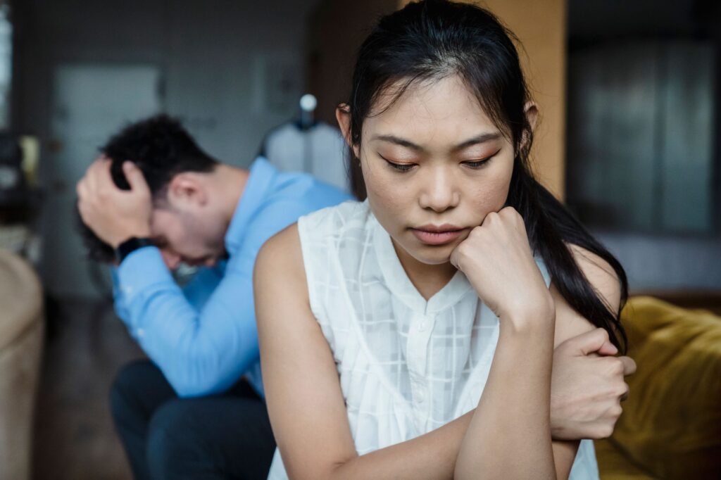 Distant couple sitting silently on opposite sides of a sofa in their living room, illustrating emotional disconnection in a relationship.